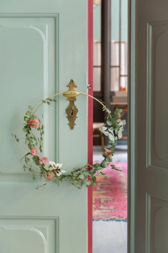 Moodbild eines goldenen Dekorationsrings umwickelt mit verschiedenen Blüten und Zweigen vor einer mintfarbenen Holztür.