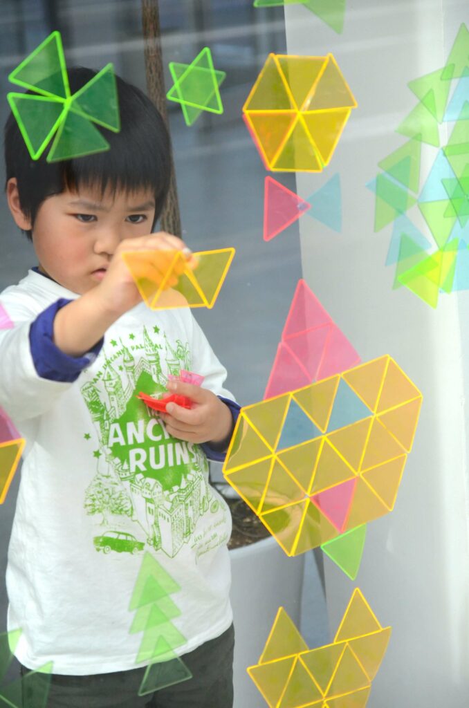 Ein Kind steht vor einem Fenster und klebt dreieckige, neonfarbene Formen in verschiedenen Kombinationen auf das Glas.